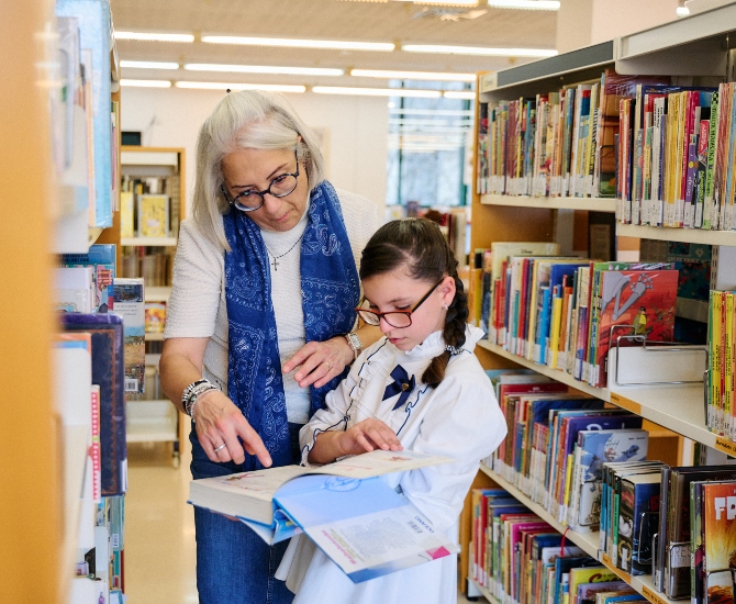 Una bibliotecària dona instruccions a una estudiant que sosté un llibre entre les mans, a una biblioteca escolar.