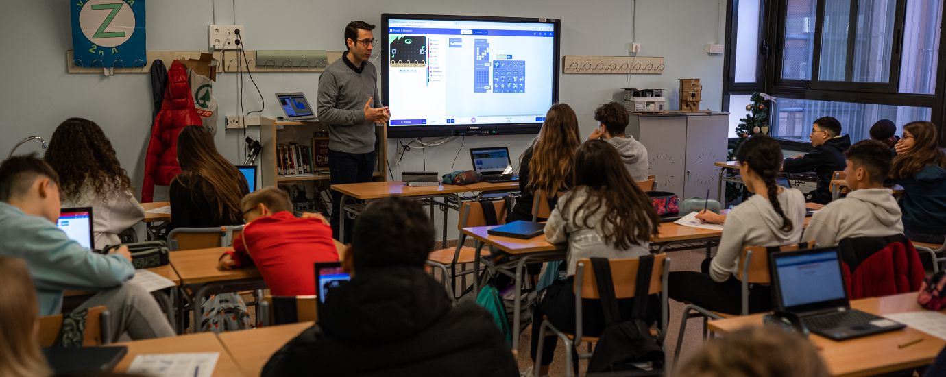 Imatge d'una aula de secundària d'un centre, on apareixen tot de joves escoltant les indicacions d'un docent, que té darrere seu una pantalla.