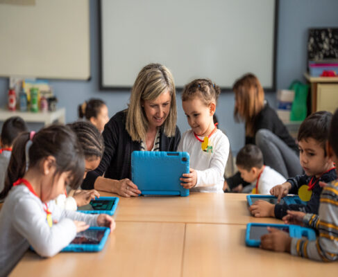 Una professora fa servir una tablet amb una alumna d'infantil. A la mateixa taula, quatre alumnes més treballan de forma individual.