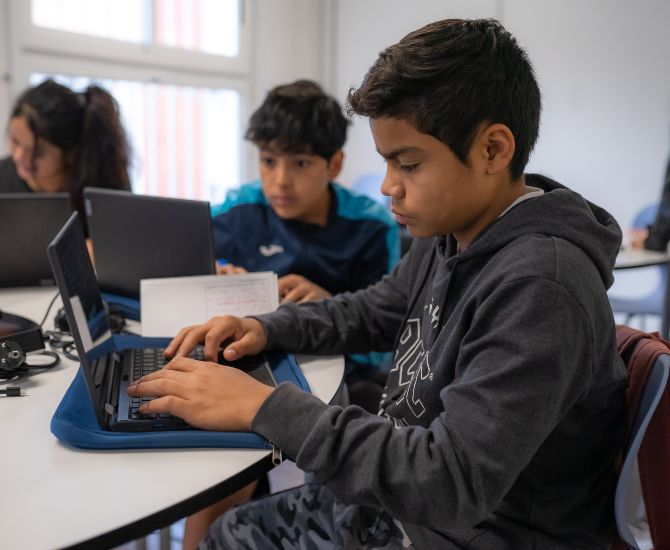 En primer pla, es veu a un estudiant escrivint a un teclat, davant de l'ordinador.