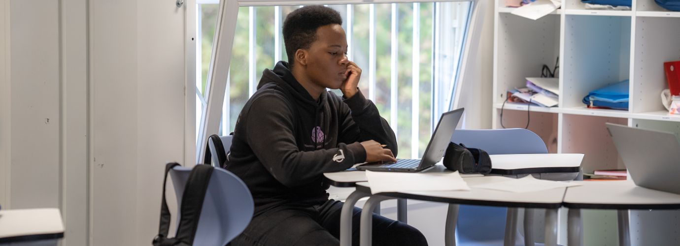 Apareix un estudiant racialitzat davant del seu ordinador, a una aula secundària, tot sol a la taula.