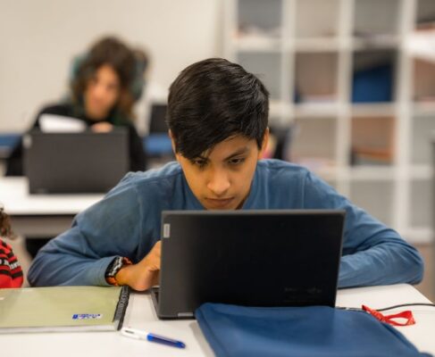 Apareix un estudiant en primer pla fent quelcom al davant d'una pantalla d'ordinador.